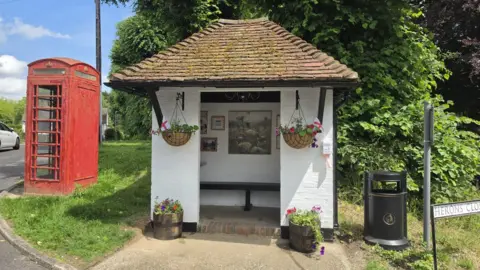 Sharon Thompson A small white bus shelter with a sloped roof. There are paintings on the wall inside and pots and hanging baskets are filled with flowers.