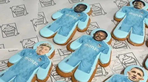 Iced biscuits shaped like football players in light blue kits are arranged on a counter.
