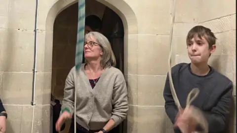 Woman in a grey cardigan and a boy in a grey jumper in an indoor church setting pulling on ropes which are attached to the ceiling.