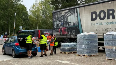 People from South East Water putting bottles of water from the water station into a blue car. There are five people in the image, and they are all wearing high-vis vests.