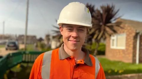 Fiona Irving / BBC A man with a white hardhat on smiles at the camera. He is wearing a high-vis orange jacket. Behind him is a residential street and a bungalow. You can make out green plastic barriers on the pavement.