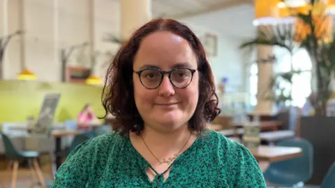 Catrin has shoulder length curly hair and wears glasses. She is wearing a green top