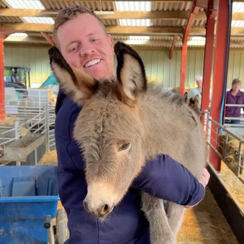 Dalscone farm Ben Best, in a navy blue jacket, lifts up the young donkey and smiles at the camera, while the foal rests his head on his arm