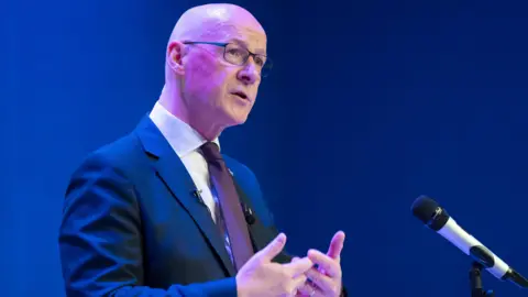 PA Media John Swinney standing on stage wearing a blue suit and a burgundy tie. There is a microphone to his right and there is a dark blue background.