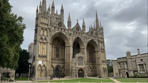 Steve Hubbard/BBC The large Peterborough Cathedral with three tall pointed arches and multiple ornate spires, set in a green lawn under a cloudy sky.