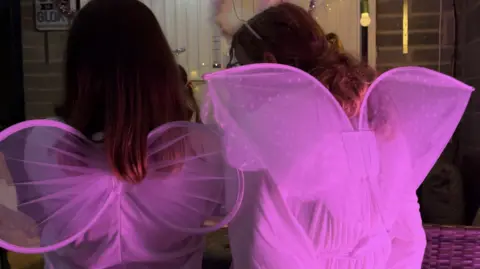 There are two girls dressed as angels with wings facing towards the nativity scene. They are lit in pink. 