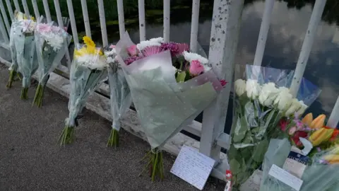 Bouquets of flower tributes are left on the floor leaning up against bridge railings. A note can be seen next to one of them.