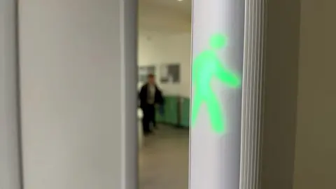 Green illuminated walking figure on a wall or panel inside a building corridor, with a blurred hallway and a person visible in the background.
