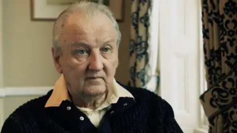 Michael Sheen Michael Gowan indossa un maglione blu con il colletto di una camicia bianca. Ha i capelli bianchi corti