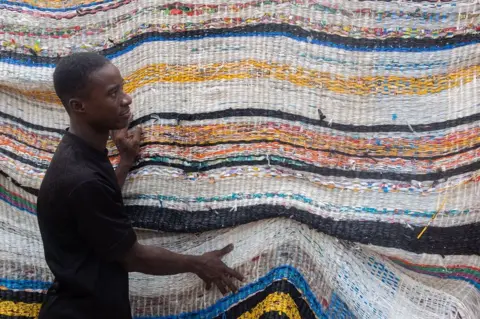 Emmanuel Adegboye / EPA A man stands next to a large woven tapestry in white, yellow blue, orange and black horizontal stripes.