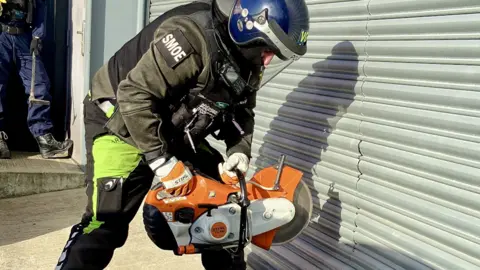 Essex Police A police officer using a saw to cut through a grey shutter. The officer is wearing gloves and a helmet. 