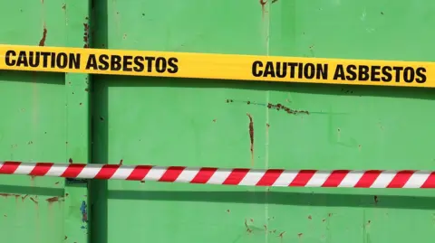 Getty Images The side of a green trailer containing absestos. Across the centre of the image runs a bright yellow warning tape printed repeatedly with the words "CAUTION ASBESTOS" in bold black capital letters.