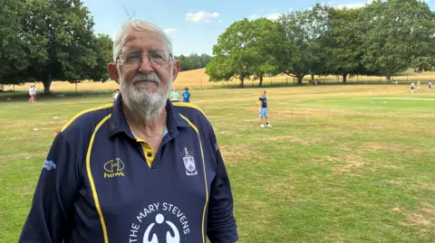 A man with a grey beard and hair is wearing glasses and on the left on the photo looking at the camera. Grass is behind him and trees are in the distance.
