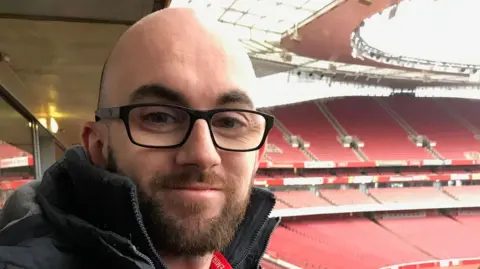Gregory Dervin smiling while on a tour of Arsenal's Emirates Stadium. He has a beard and shaved head and is wearing glasses and a black coat. Behind him are row upon row of empty red seats.