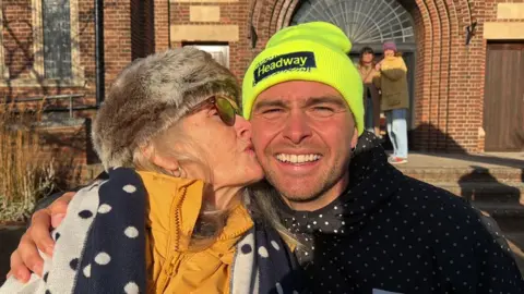 A close-up of Penny and her son Sam King, who has his right arm around her, outside with a brick-built church behind them and she is kissing his cheek. She is wearing a fur trimmed hat, dark glasses and a yellow puffer jacket with a blue and white scarf and has long fair hair. He is wearing a bright yellow beanie hat and a black windcheater with white dots. 