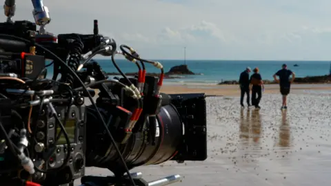 BBC Close-up of a professional film camera positioned on a beach, sharply focused against a blurred coastal background. Three people walk near the water's edge across wet sand reflecting the sky, while calm blue water and a small rocky structure sit along the horizon.