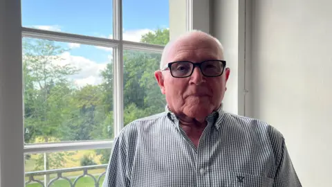 Roy is wearing a short-sleeved checkered shirt, stands indoors near a multi-paned window. Outside the window, trees, greenery, and parked cars are visible