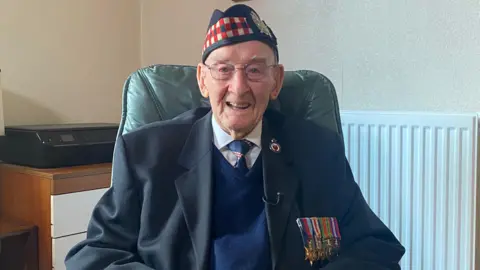 A man is sat on a green chair smiling at the camera. He is wearing a white shirt with a navy tie, jumper and blazer. He has a navy hat on with a red, white and black stripped pattern along the front. He has a row of medals displayed on his blazer.