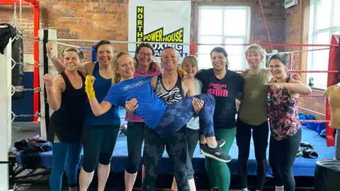 Nine women in gym gear stand in front of a boxing ring.