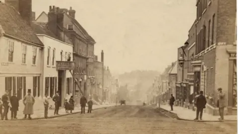 St Albans Museums A sepia image from about 1900 looking down Holywell Hill in St Albans. There are buildings on each side of the road and one cart can be seen on the road down the hill. There are people on the pavements in Victorian dress. 