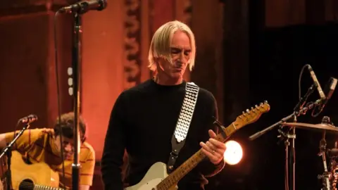 Paul Weller playing a guitar, with blond hair in a centre parting, looking down at the guitar. In the background is another guitarist and the edge of a drumkit.