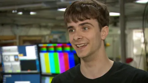 A man is stood looking past the camera in a black t-shirt. There is a computer screen in the background with multi-colours on it.