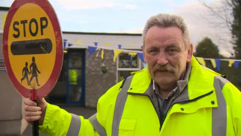 Brendan McLenaghan has grey hair and a moustache. He is wearing a hi-vis jacket and holding a circular sign that says stop.