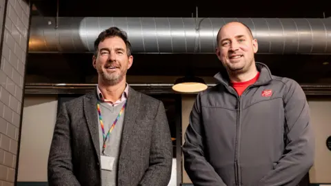 Dressed in jackets Stephen Underwood from Jersey Electric and Captain Richard Nunn from the Salvation Army stand in front of a large silver coloured pipe and café tables. The cafe has white tiles on the walls and dark green paint.