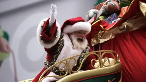 Getty Images An image showing Santa Claus at the Macy's Parade in New York in 2010. He is wearing his traditional red and white costume and hat, and is gesturing cheerfully from a sleigh at the crowd. Beside him is a giant bag full of wrapped toys, an American football and a soccer ball. 