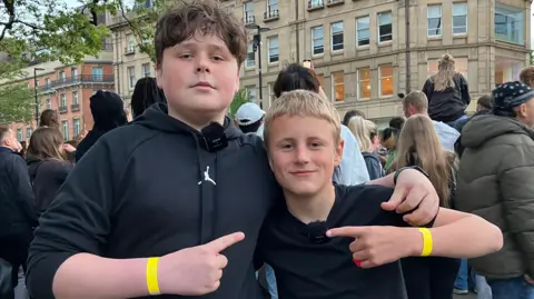 Two young boys stand in front of a crowd. They are pointing at each other, each of them wear a yellow wristband.