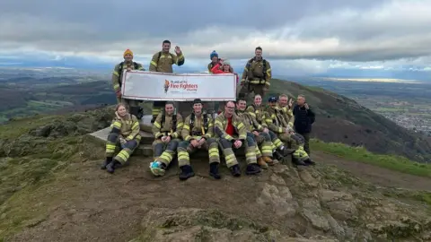 Tom Brooks-Simon A group of people in firefighting uniform photographed at a summit trig point, carrying a Fire Fighters Charity banner.