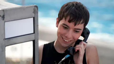 Ellis Howard as Byron in What It Feels Like For A Girl, using a pay phone and smiling.