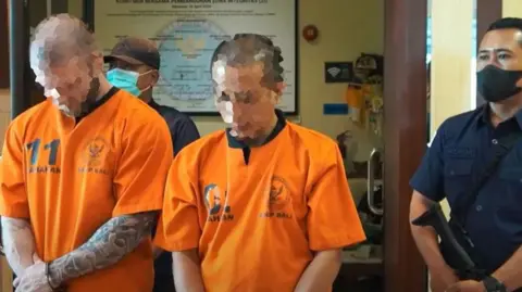 BNNP Bali Two men in bright orange detention tops stand next to an officer holding a gun.