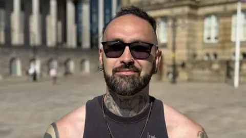 A close-up of a man with dark hair and a beard, wearing dark sunglasses. he has a large tattoo on his neck and arms.