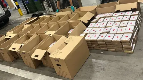 Europol Cardboard boxes containing parcels with cocaine have been lined up after a police raid. Some of the parcels have been placed next to the boxes. They are wrapped in brown tape and have an ace of diamonds on the top. 