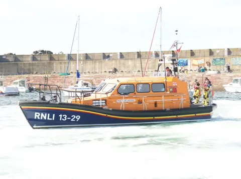 Robbie Bald A lifeboat in orange and blue sails through the water  with a pier or harbour behind it