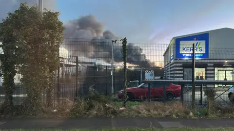Plumes of grey smoke are emerging from an industrial estate, in the distance. There is a building with the sign Kerr's Tyres at the right of the image. A red car is parked outside