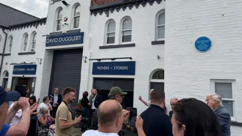 A crowd gathered outside a building with a circular blue plaque on the wall. The building on the left has a dark blue sign above the door that reads AUCTIONEERS - VALUERS, DAVID DUGGLEBY, MOVERS - STORERS