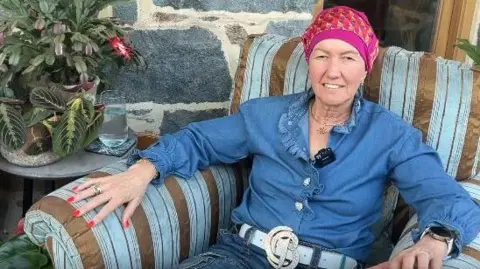 Dressed in a denim blue top with a pink headscarf Catherine Best has a mike  clipped to her lapel as she looks straight at the camera for an interview. 