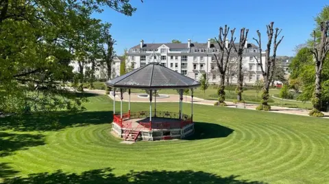 A bandstand is surrounded by freshly mown circular lawns in Dane John Gardens, with houses and trees in the background.