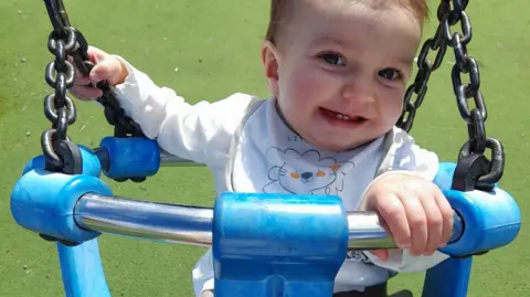 A baby boy in a blue swing, smiling. He is holding the metal side of the swing and wears a white top and bib with a lion on.