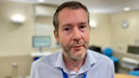 Mark Norman BBC Dr Mike Bedford in a blue shirt standing in a hospital room with computers in the background.