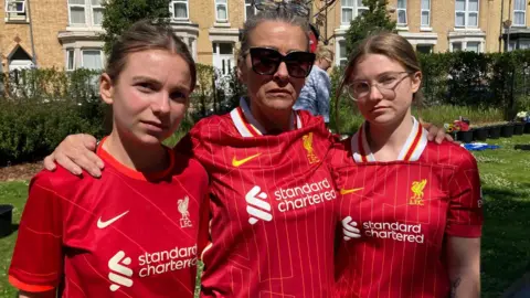 Marc Gaier/BBC Andrea Molyneux, who is wearing large sunglasses, stands with her arms around the shoulders of her daughters either side of her. All three women are wearing Liverpool kits and have sombre expressions.