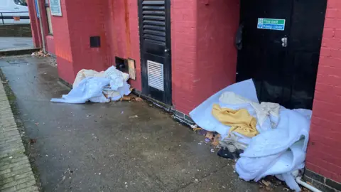 A pathway with white mattresses bundled up with various cloths on them showing rough sleeping on a street in Coventry outside a red brick-walled building.