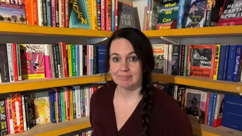 A woman with dark hair and a dark top standing in front of a corner book case. 