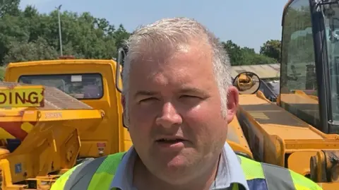 A man in a high‑visibility vest stands in front of yellow construction machinery outdoors.
