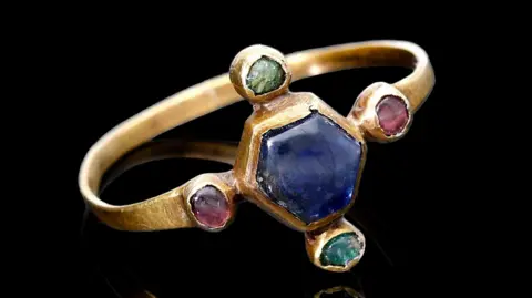 A gold ring, with a blue sapphire in the middle, then two green emeralds and two garnets either side, sitting on its side on a black backing.