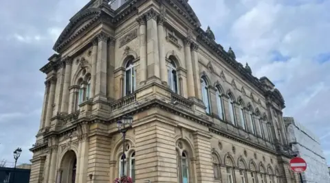 Huddersfield Town Hall, which is a Grade II-listed building, with clouds gathering overhead. The back right hand side of the building has building work taking place, with scaffolding in place.