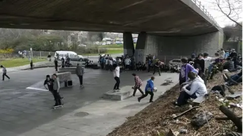 Gateshead Council A crowd of young skateboarders at the Five Bridges skatepark. Several are rolling around on their boards. Dozens more are sitting around the edge of the park looking at the others. The park is under the concrete Gateshead flyover.