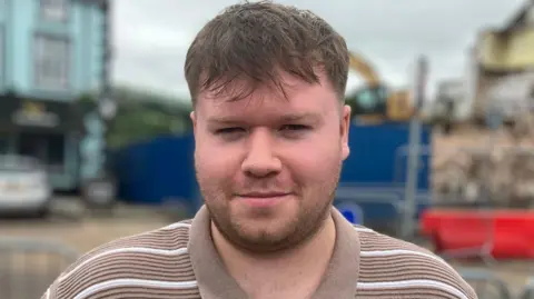 Ciaran Dallat has long brown hair and stubble. He is standing in front of the demolished site and a blurred digger is visible in the background